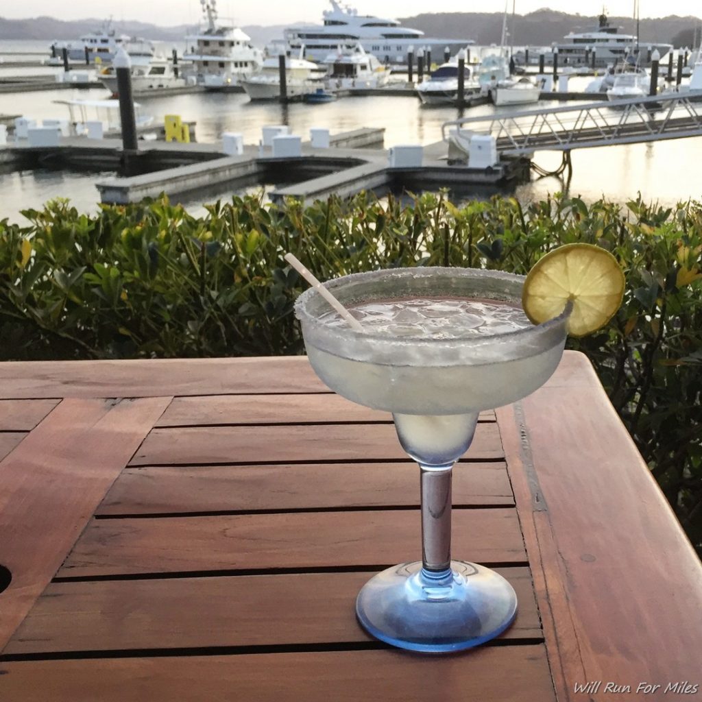 a glass of liquid on a table with a dock and boats in the background