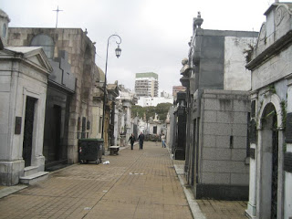 a group of people walking down a street with La Recoleta Cemetery in the background