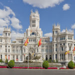 a large white building with a fountain and flags with Plaza de Cibeles in the background