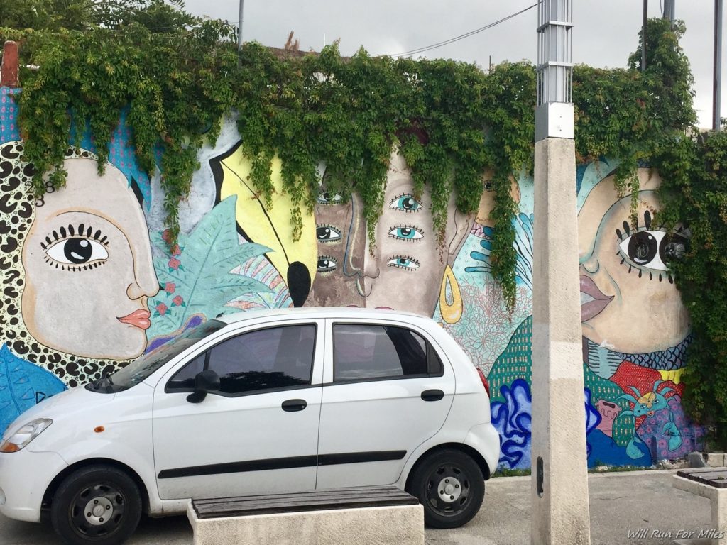 a white car parked in front of a wall with a painting of faces