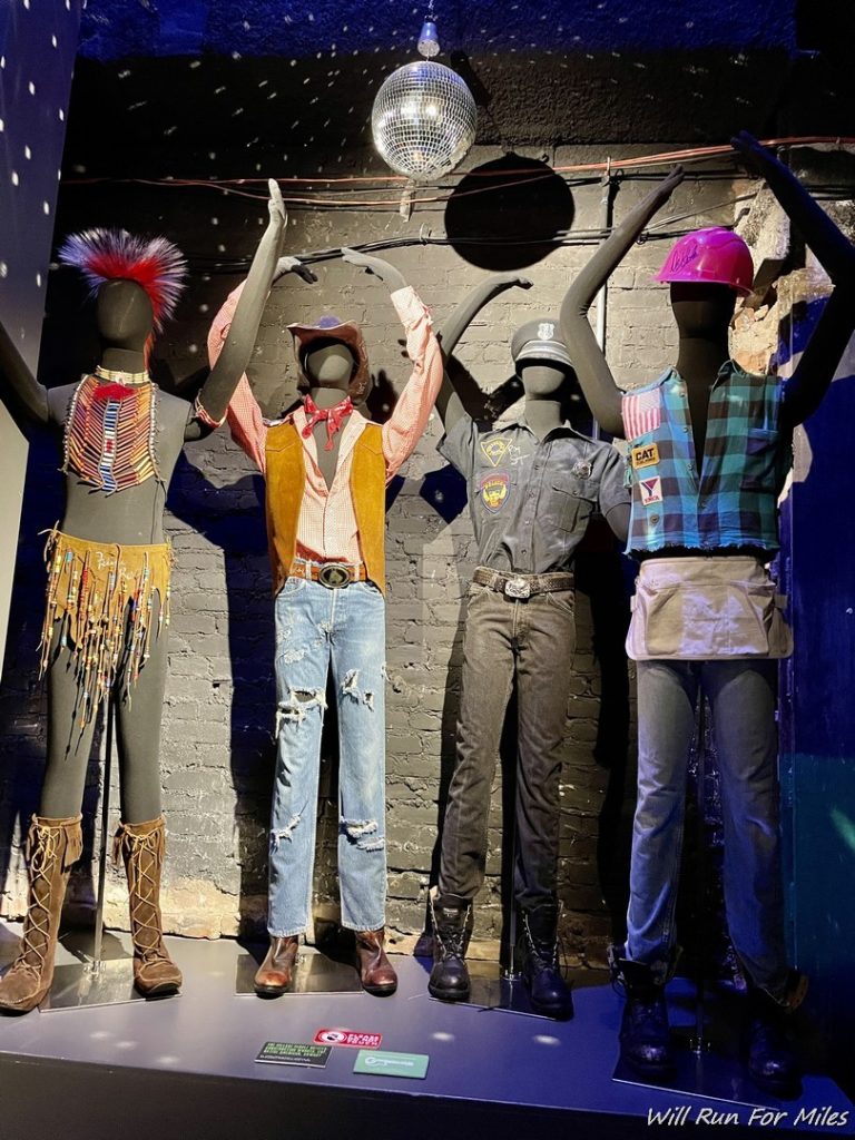 mannequins with arms raised in a room with a black wall and a ball and a disco ball in the background