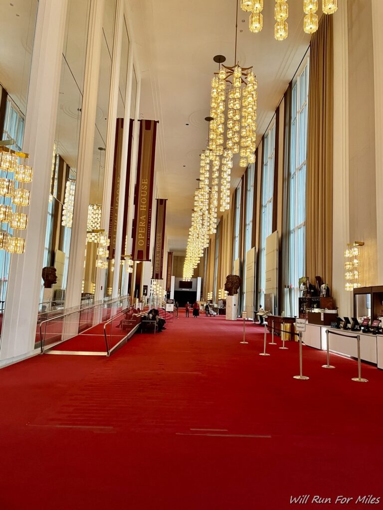 a long hallway with red carpet and chandeliers with John F. Kennedy Center for the Performing Arts in the background