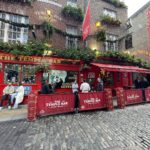 a red bar with people sitting in front of it with Temple Bar, Dublin in the background