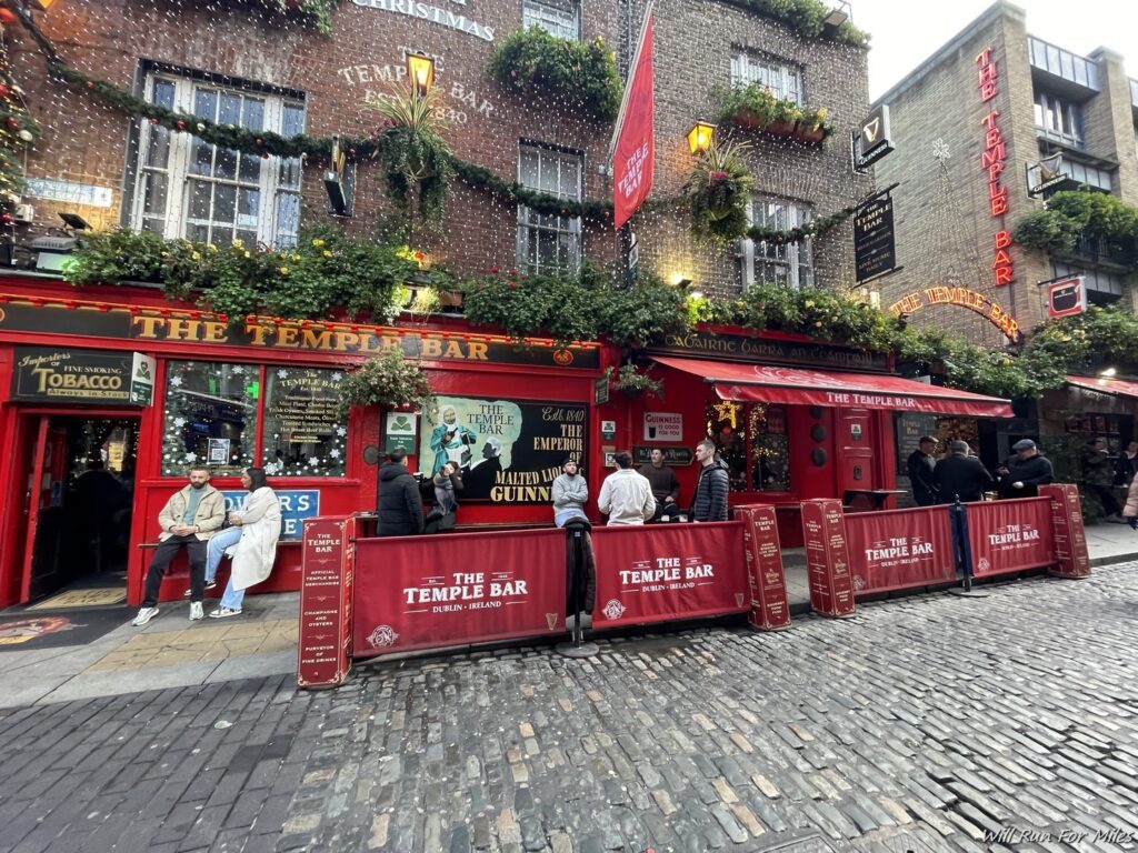 a red bar with people sitting in front of it with Temple Bar, Dublin in the background