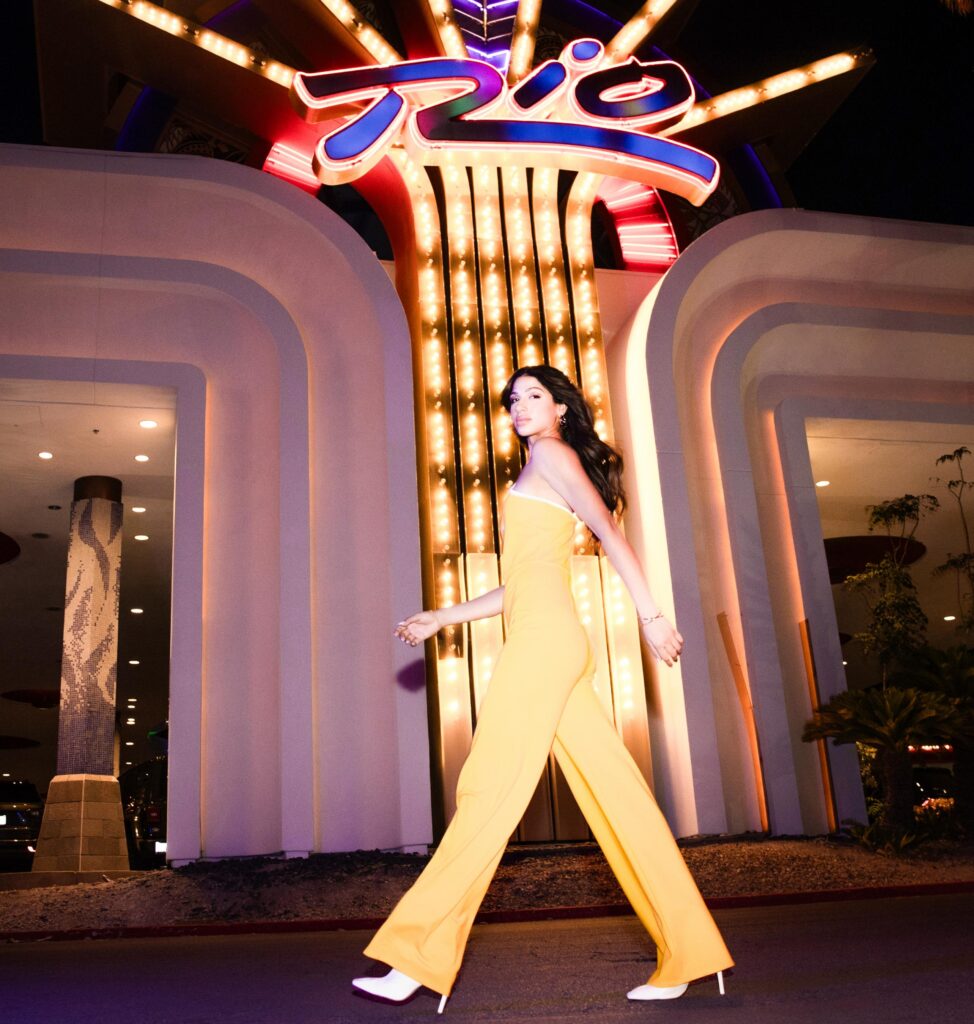 a woman in yellow jumpsuit walking in front of a neon sign